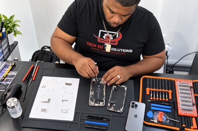 A TechEd and Solutions technician working on the internals of an opened iPhone, with the screws laid out on a labelled repair mat